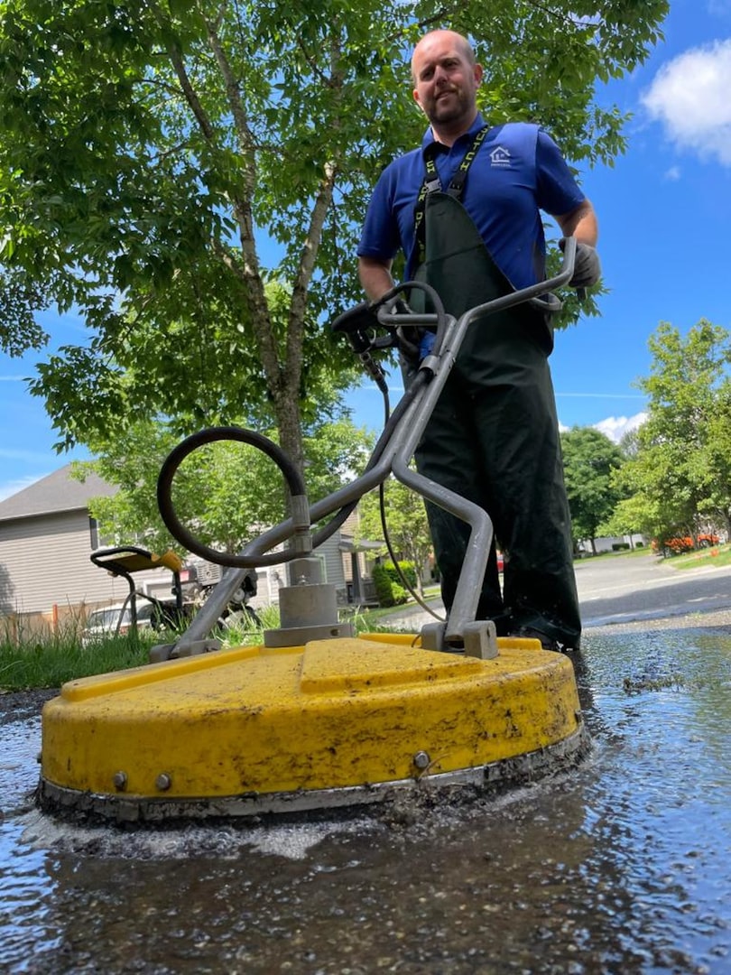 A person wearing a gray cap, gray long-sleeve shirt, blue overalls, and yellow rubber boots is power washing a black garage door. There is a hose on the ground, and the surface appears wet. A small tree is visible nearby.